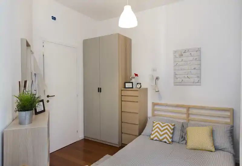 Camera da letto luminosa e ben curata con struttura del letto in legno, cuscini decorativi e mobili contenitore coordinati; pavimento in legno caldo e pareti neutre per un'atmosfera tranquilla.