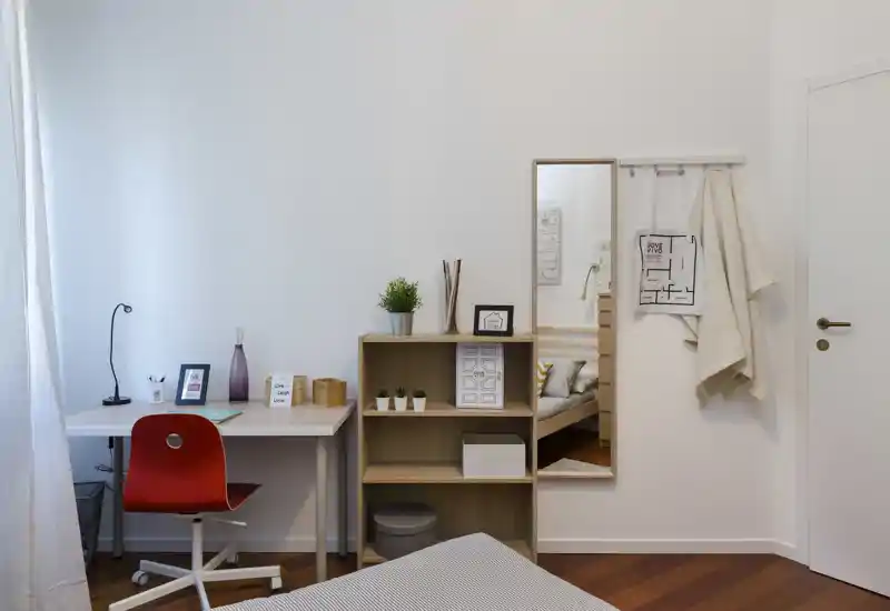 Angolo camera luminoso e ordinato con scrivania, scaffale e letto parzialmente visibile in primo piano — ben illuminato e arredamento semplice e confortevole.