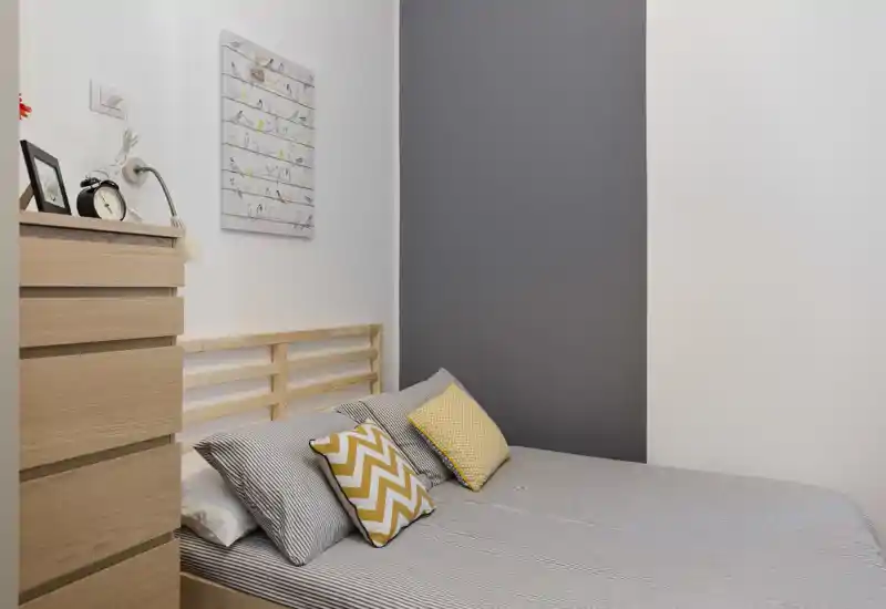 Camera da letto compatta e moderna con testiera in legno, biancheria a righe e cuscini decorativi gialli; ambiente minimale e ben curato, adatto a letto singolo o matrimoniale.