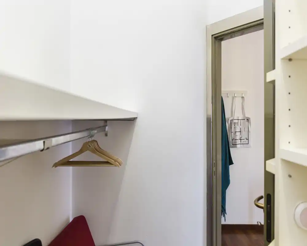 Pequeña zona de entrada/armario con barra para colgar y perchas, paredes blancas y una puerta que conduce a una habitación con suelo de madera.