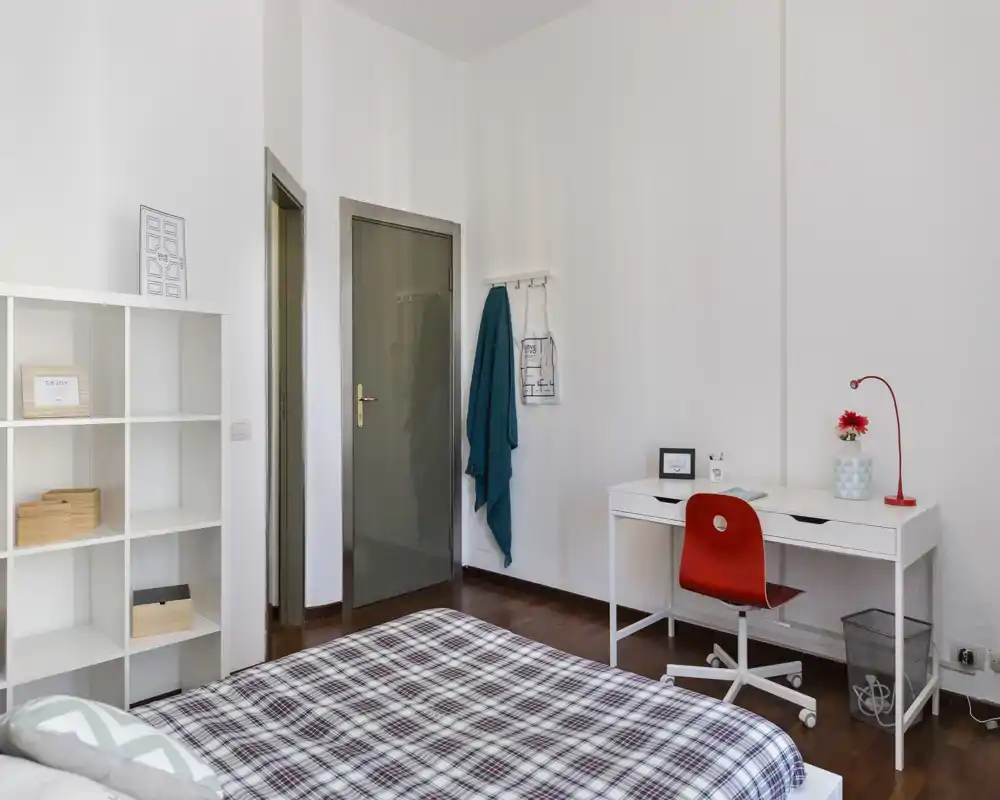Dormitorio luminoso y ordenado con colcha de cuadros, estantería y escritorio blanco con silla roja: un espacio moderno ideal para descansar y trabajar.