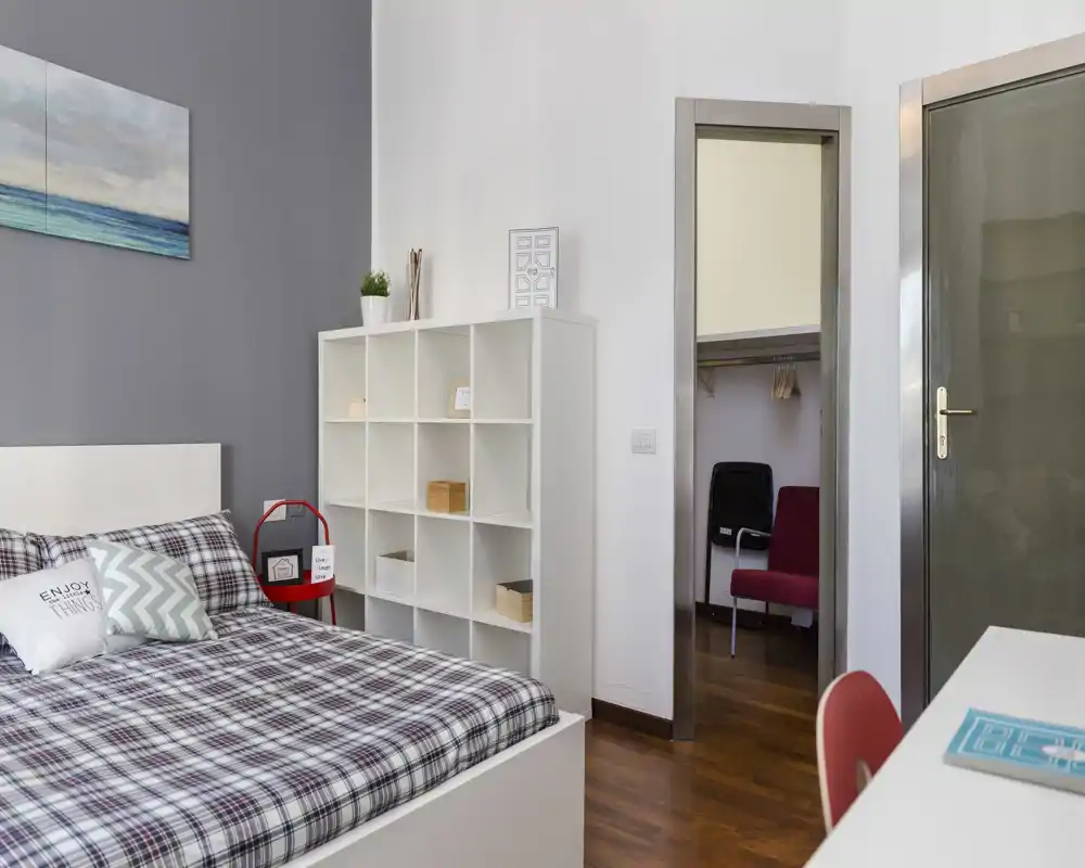 Dormitorio luminoso y bien decorado con una cama grande de cuadros, cojines decorativos y una estantería modular — un espacio contemporáneo y confortable ideal para relajarse.