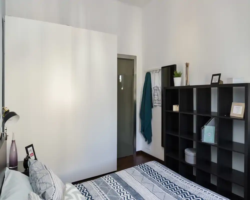 Camera da letto luminosa e ordinata con letto matrimoniale dal motivo geometrico, ampio armadio bianco e scaffalatura a vista—linee pulite e luce soffusa creano un ambiente accogliente.