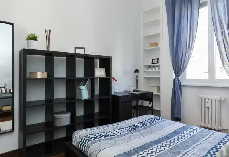 Camera da letto luminosa e ordinata con letto matrimoniale con copriletto blu a motivi, una libreria a giorno e una piccola scrivania vicino alla finestra che lascia entrare luce naturale.