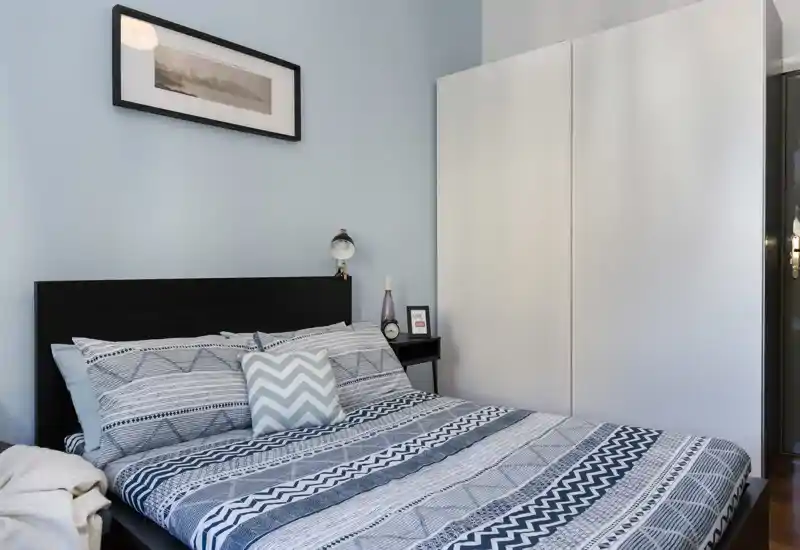 Camera da letto luminosa e ordinata con letto matrimoniale a fantasia, lampada da comodino e grande armadio bianco — spazio moderno e accogliente ideale per riposare.
