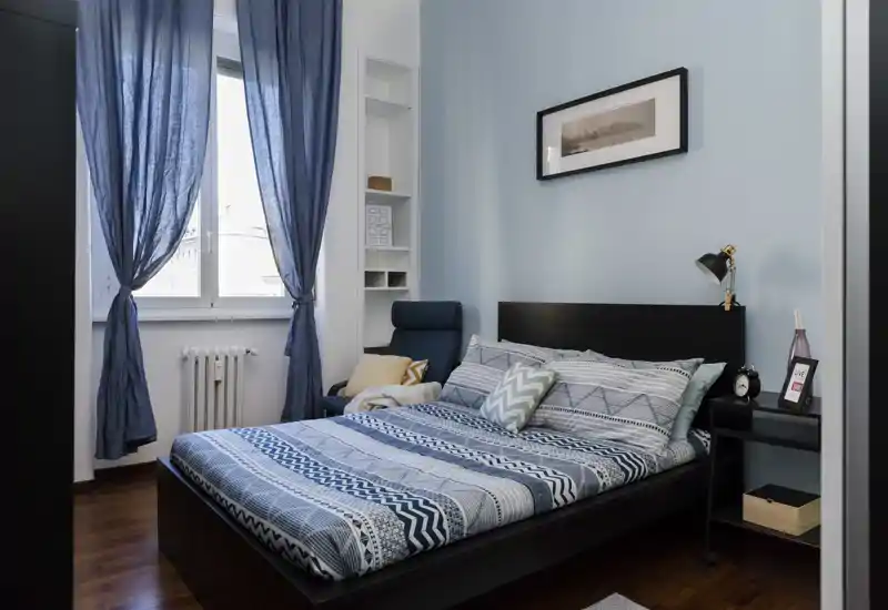 Camera da letto luminosa e ben allestita con letto matrimoniale con biancheria blu a fantasia, comodino con lampada e poltrona vicino alla finestra — perfetta per annunci.