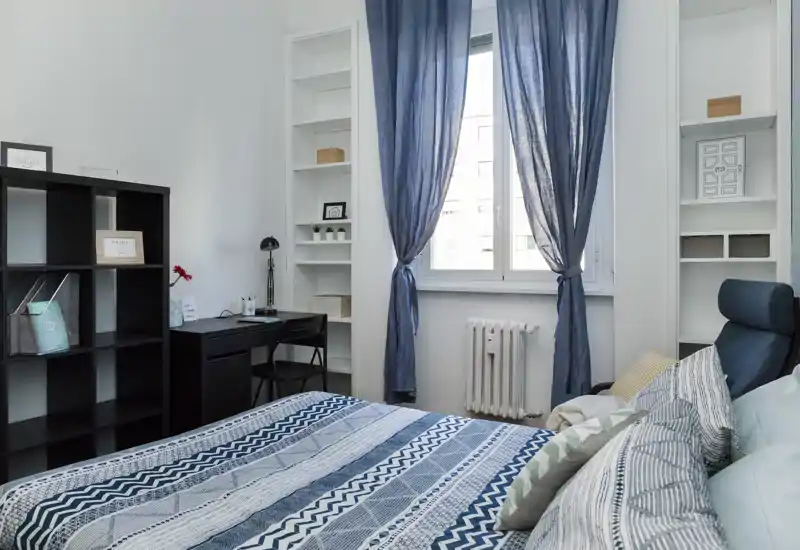 Camera da letto luminosa e ben arredata con ampio letto con biancheria blu a motivi, poltrona per la lettura, scrivania e scaffalature accanto alla finestra che lascia entrare luce naturale.