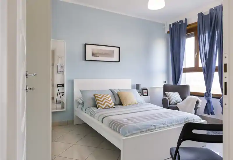 Dormitorio luminoso y bien decorado con cama de estructura blanca, cojines estampados y paredes azul claro; la luz natural de la ventana aporta una atmósfera tranquila y acogedora.
