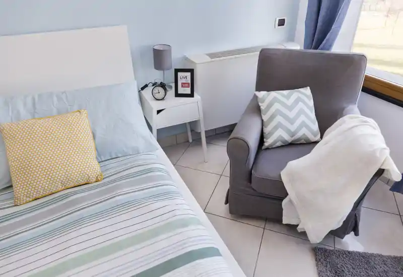 Rincón de dormitorio luminoso y bien arreglado con cama a rayas, mesita de noche y un sillón cómodo junto a la ventana — ideal para una estancia acogedora.