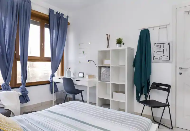 Dormitorio luminoso y bien decorado con cama a rayas en primer plano, escritorio junto a una gran ventana y estantería elegante; la luz natural y los textiles crean un ambiente acogedor.