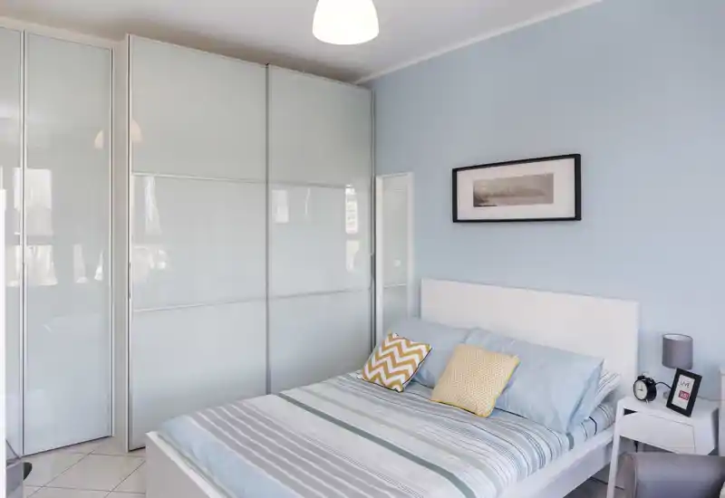 Dormitorio moderno y luminoso con cama blanca, cojines estampados y un gran armario empotrado con puertas correderas traslúcidas. Paredes azul claro y ropa de cama coordinada crean un ambiente sereno y acogedor.