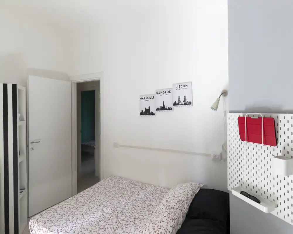 Dormitorio luminoso y ordenado con cama doble vestida con ropa de cama estampada, estantería minimalista y una pequeña lámpara de noche. Paredes blancas y decoración sencilla crean un ambiente tranquilo y fresco.