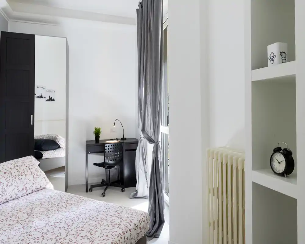 Dormitorio luminoso y minimalista con edredón estampado, escritorio junto a la ventana y armario con espejo — ordenado y bien iluminado, ideal para una persona o estudiantes.