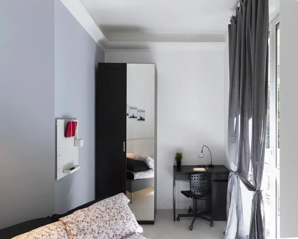 Dormitorio moderno y luminoso con cama doble, armario con espejo y un escritorio compacto junto a la ventana; tonos neutros y ropa de cama con estampado suave que aportan calma.