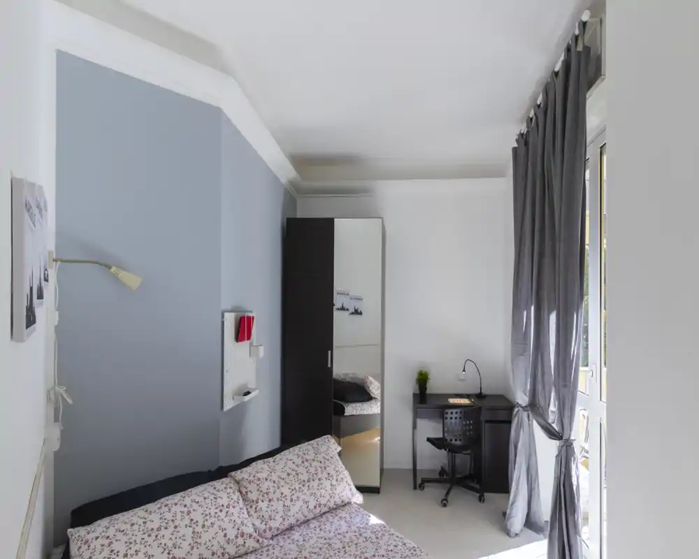 Dormitorio luminoso y ordenado con edredón estampado, armario con puerta espejo y un pequeño escritorio junto a la ventana que aporta luz natural.