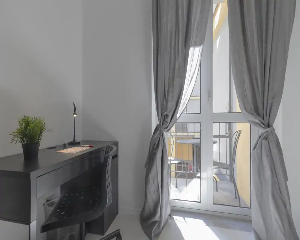 Rincón luminoso de salón con pequeño escritorio y puerta al balcón. El espacio incluye una zona de trabajo ordenada con planta, lámpara de escritorio y cortinas largas que se abren a una pequeña área exterior con sillas.