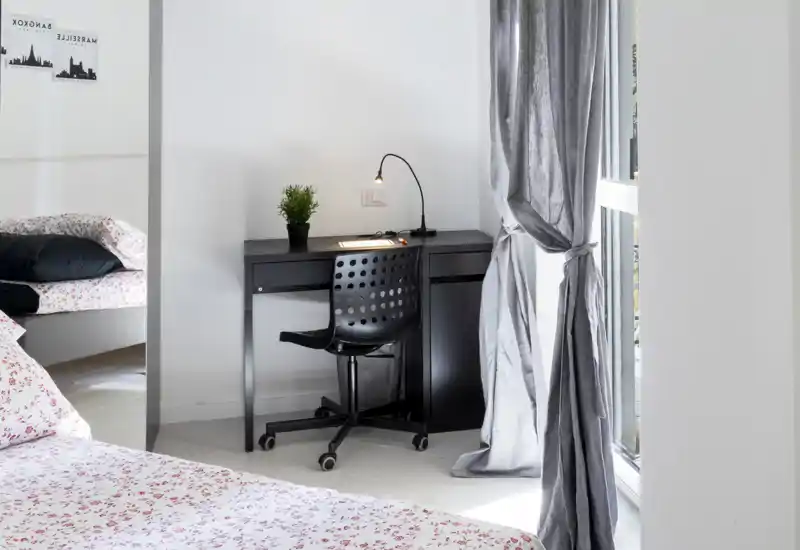 Angolo camera da letto luminoso e moderno con letto matrimoniale in primo piano, biancheria a fantasia e una scrivania nera compatta con lampada vicino a una grande finestra.