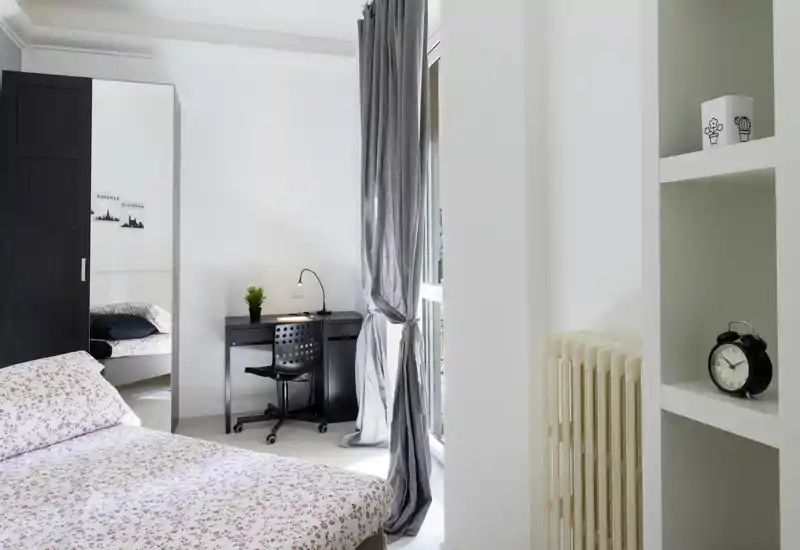 Camera da letto luminosa e minimal con copriletto a fantasia, scrivania vicino alla finestra e armadio con specchio — ordinata e ben illuminata, ideale per una persona o studenti.