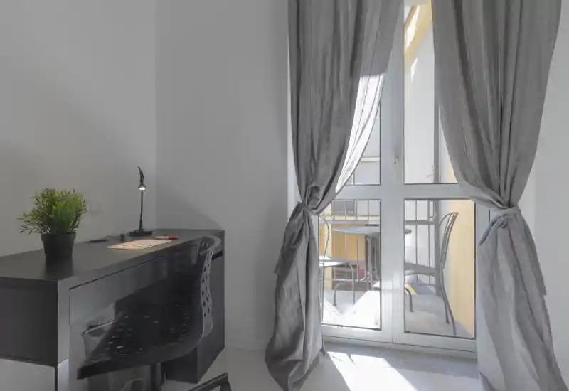 Zona giorno luminosa e minimale con una scrivania accanto alla porta del balcone; la luce naturale morbida inonda la stanza e le tende incorniciano un'area esterna con sedute.