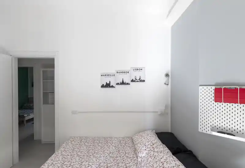Camera da letto luminosa e minimal con biancheria a fantasia, piccole stampe a parete e una palette bianca che enfatizza spazio e tranquillità.