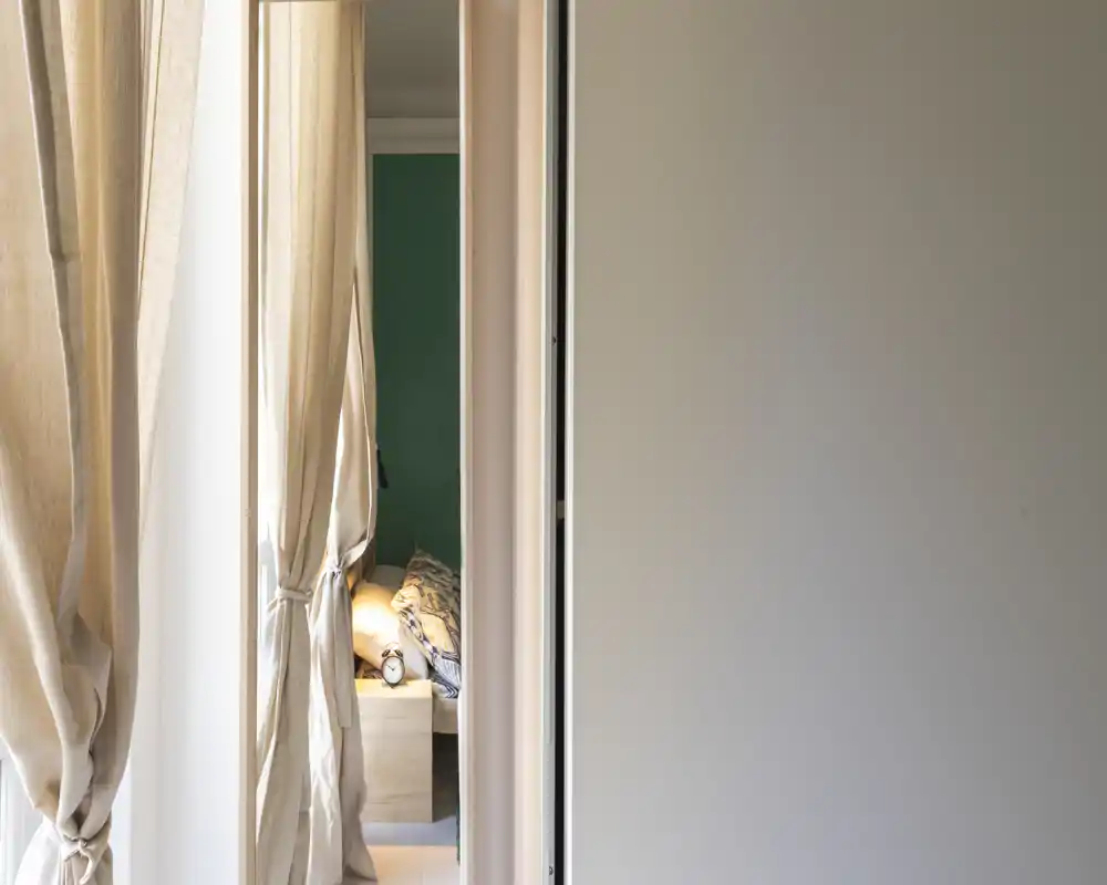 Chambre doucement éclairée aperçue à travers une porte avec rideaux noués ; on voit une table de chevet, des oreillers et une partie du matelas, créant une ambiance chaleureuse.