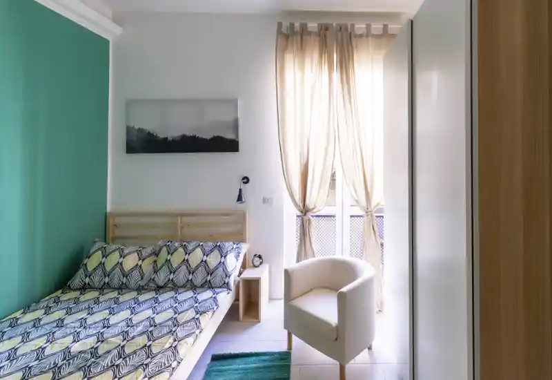 Dormitorio moderno y luminoso con ropa de cama estampada, estructura de cama de madera clara, un sillón acogedor y cortinas con luz natural — ideal para un anuncio.