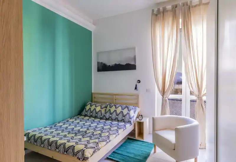 Dormitorio luminoso y bien decorado con cama doble frente a una pared turquesa, luz natural desde el balcón, sillón y mesita — acogedor y listo para huéspedes.