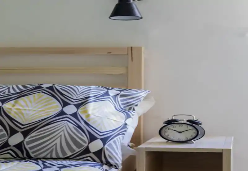 Rincón de dormitorio luminoso y moderno con almohada estampada, cabecero de madera sencillo y mesita con un despertador clásico. La imagen resalta una zona de descanso acogedora y bien iluminada, ideal para un anuncio.