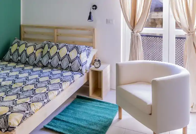 Dormitorio moderno y luminoso con cama de madera, ropa de cama estampada, mesita cubo y un sillón junto a la ventana soleada.