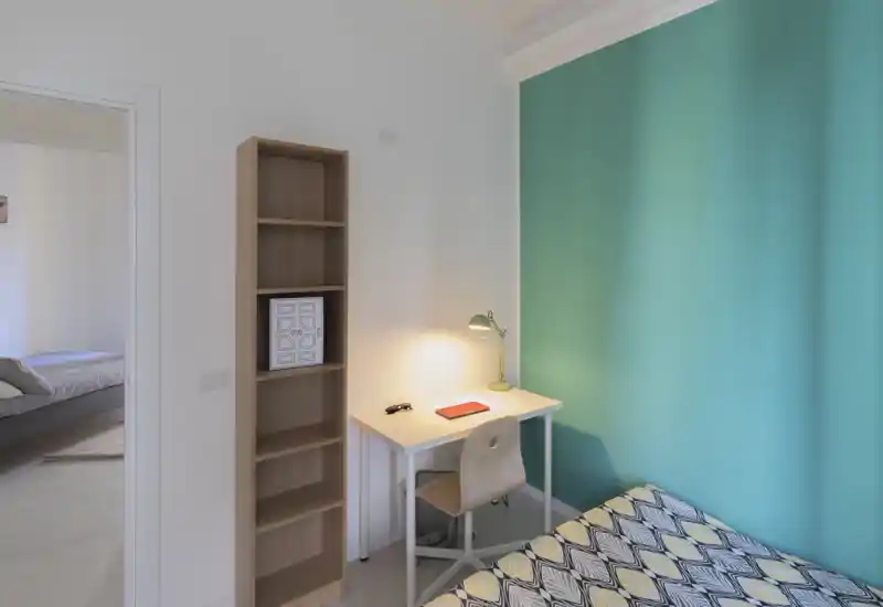 Small, bright bedroom corner with a patterned single bed, compact desk and a teal accent wall. The space is well lit and furnished for a comfortable single stay or student rental.