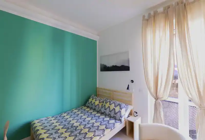 Dormitorio luminoso y acogedor con cama doble estampada, luz natural desde el balcón y pared de acento color turquesa.