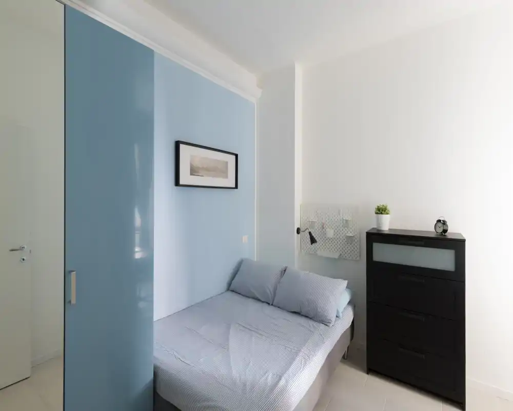 Compact, bright bedroom with a neatly made double bed, soft blue accents and a tall black chest of drawers. Minimalist decor and natural light create a calm atmosphere.