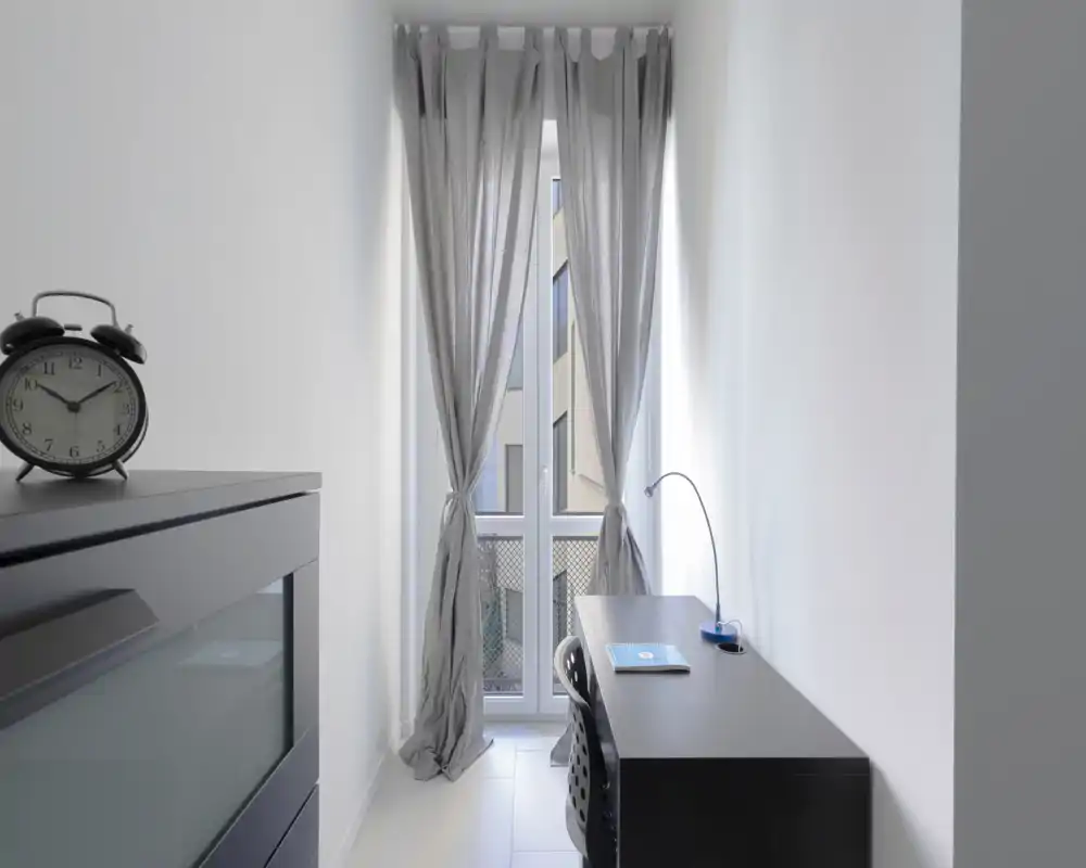 Narrow living / work corner with a tall window dressed by light curtains; a desk and chair create a bright, minimal workspace.