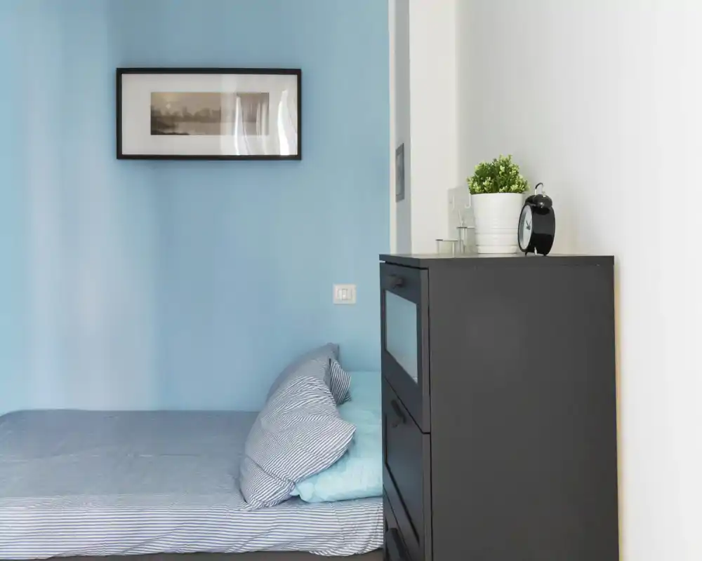 Compact bedroom corner with a partially visible single bed dressed in striped linens, soft blue walls and a dark chest of drawers topped with a plant and alarm clock.