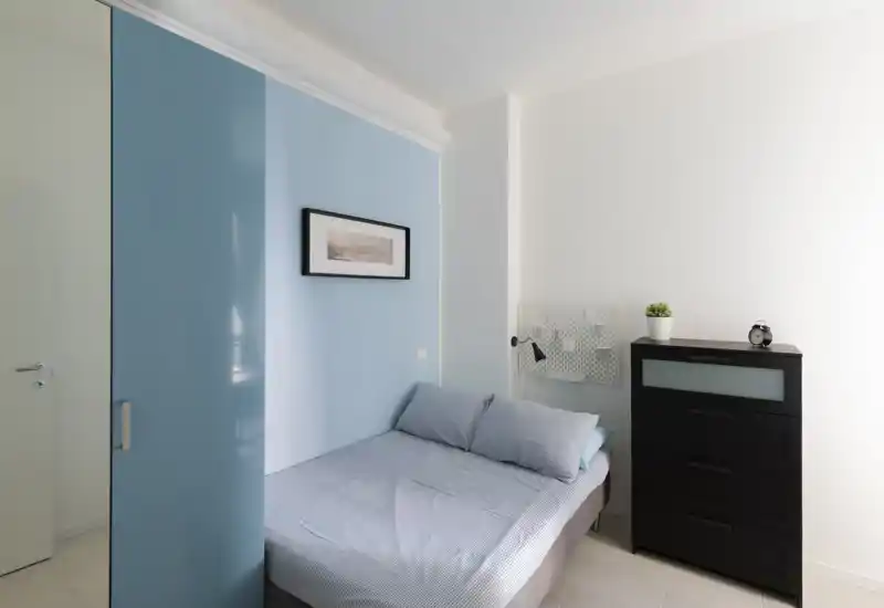 Dormitorio compacto y luminoso con cama doble bien hecha, armario azul claro y cómoda oscura — moderno y ordenado.