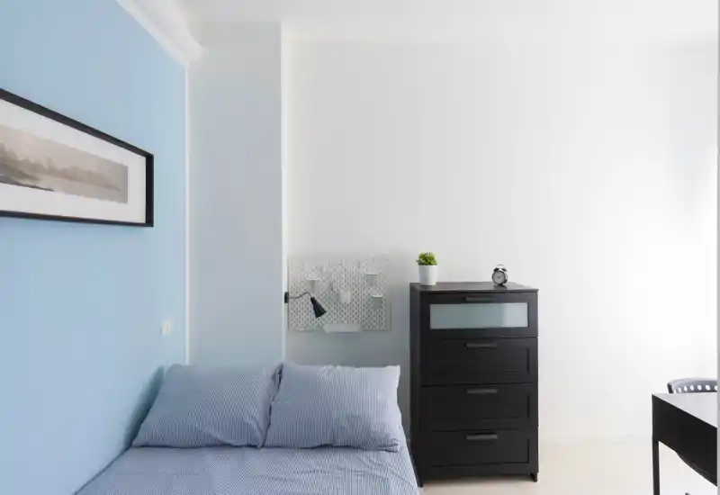 Dormitorio luminoso y minimalista con una cama individual bien hecha contra una pared azul claro, una cómoda negra y una decoración de pared sencilla — espacioso y bien iluminado.