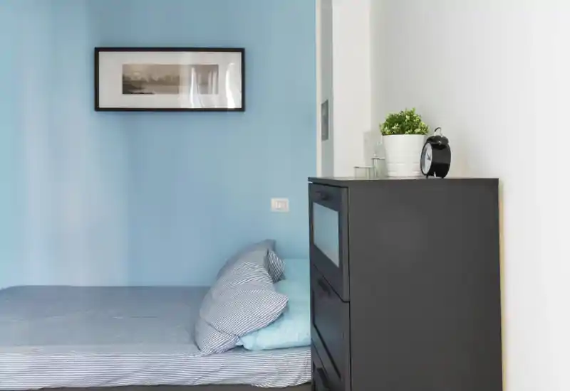 Dormitorio compacto y luminoso con una cama individual bien hecha, ropa de cama a rayas y una cómoda oscura; las paredes azul claro crean un ambiente tranquilo.