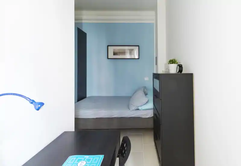 Pequeña habitación moderna con cama doble bien hecha frente a una pared azul, cómoda alta negra y una zona de trabajo compacta en primer plano. Decoración mínima y luminosa que aporta un ambiente tranquilo y funcional, ideal para alquileres cortos.
