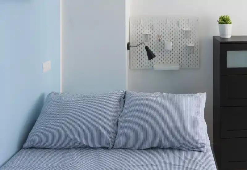 Dormitorio luminoso y minimalista con cama doble de rayas, lámpara de lectura y pequeño armario; tonos azul y blanco que provocan sensación de tranquilidad.