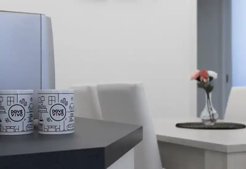 Angolo cucina/pranzo moderno con piano scuro in primo piano, due tazze brandizzate, sedie bianche e un piccolo tavolo con un vaso di fiori — pulito, luminoso e dallo stile essenziale.
