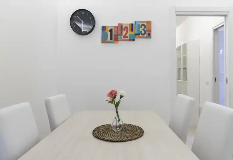 Zona pranzo luminosa e minimale con tavolo in legno chiaro, quattro sedie imbottite bianche e un semplice vaso di vetro al centro; stile pulito e contemporaneo adatto per annunci.