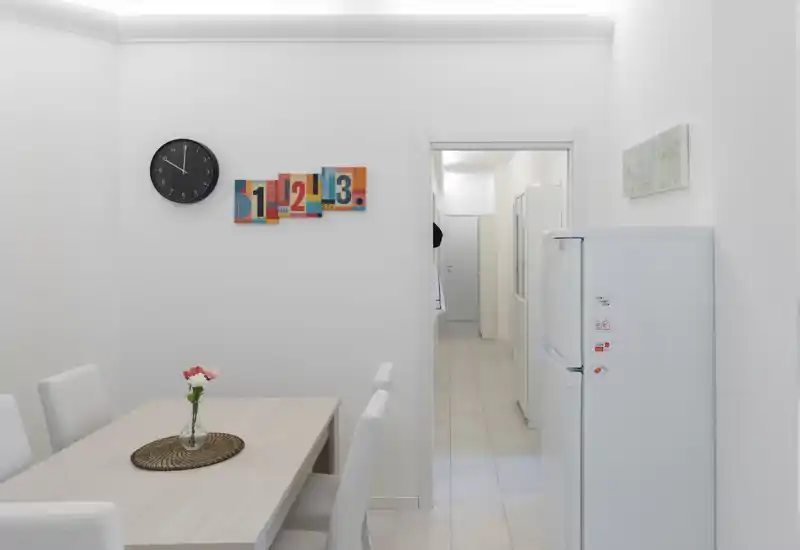 Zona pranzo luminosa e minimal con tavolo in legno chiaro, sedie bianche e un piccolo vaso di fiori; lo spazio si apre su un corridoio e sul lato destro si vede un frigorifero.