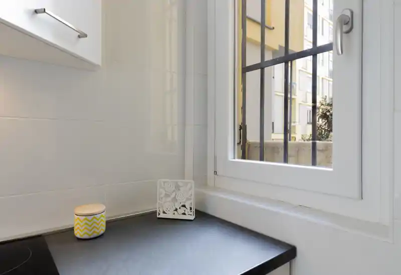 Angolo cucina compatto e luminoso con piano scuro, pareti piastrellate bianche e una finestra che lascia entrare luce naturale. Un vasetto decorativo e un pannello aggiungono un tocco domestico ordinato.
