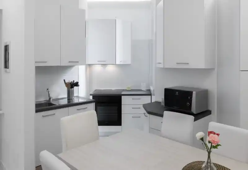 Cucina compatta moderna e luminosa con mobili bianchi, forno incassato, microonde e piccolo tavolo da pranzo; stile minimal con fiori freschi.
