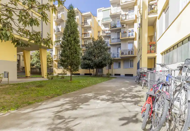 Cortile condominiale soleggiato con parcheggio per biciclette, alberi maturi e balconi degli appartamenti affacciati su un piccolo prato — ideale per mostrare lo spazio esterno condiviso.