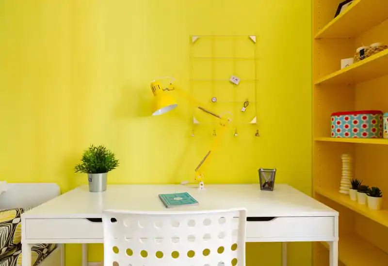 Angolo scrivania moderno e luminoso con tavolo bianco, sedia forata e lampada da tavolo gialla regolabile su pareti gialle — arredato con una piccola pianta e mensole decorative.