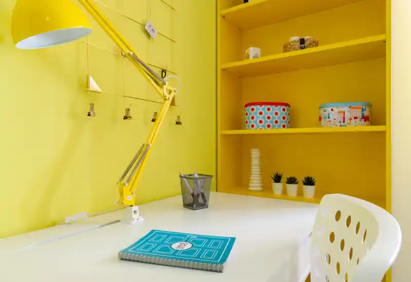 Angolo studio luminoso e moderno con scrivania bianca, lampada regolabile gialla e scaffalatura gialla con contenitori decorativi e piccole piante.