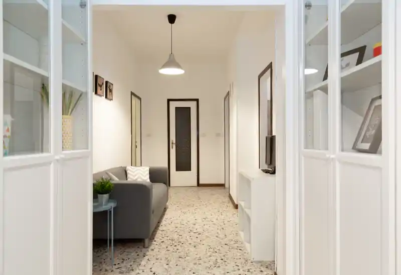 Soggiorno luminoso e minimale con divano grigio, cuscini decorativi e scaffalature; pavimento in terrazzo e lampadario a sospensione creano un'atmosfera moderna e ariosa.