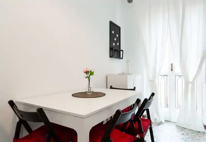 Zona pranzo minimal e luminosa con tavolo bianco, quattro sedie nere con cuscini rossi, un piccolo vaso con fiori e tende leggere che lasciano entrare la luce naturale.