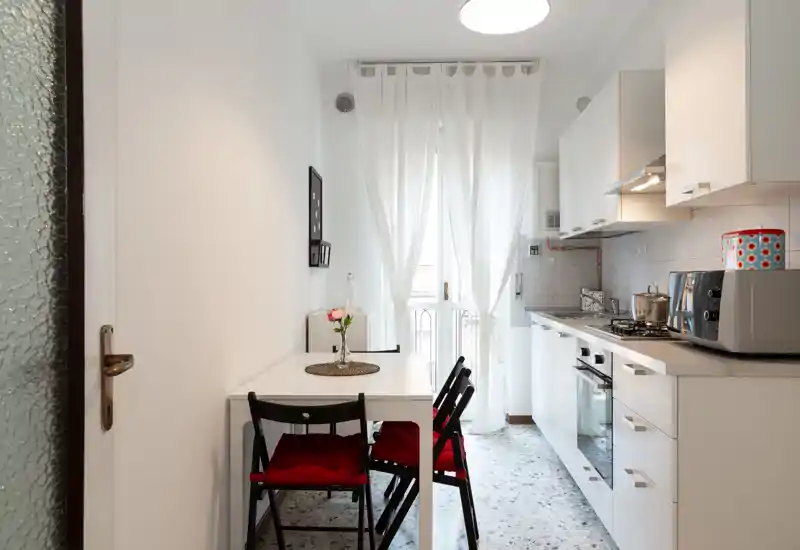 Cucina moderna compatta con pensili bianchi, piano cottura a gas e forno incassato, tavolo da pranzo piccolo con sedie dai cuscini rossi e tende leggere che diffondono luce naturale.